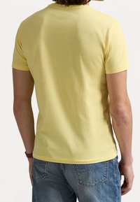 T-shirt giallo a maniche corte visto da dietro, realizzato in morbido cotone, con scollo rotondo e vestibilità rilassata, abbinato a jeans blu.