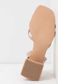 Sandale beige à talon bloc, vue du côté de la semelle, appuyée contre un mur d’angle blanc et une surface de sol.