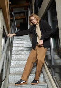 Giacca in pile nera, camicia beige, pantaloni larghi marroni e sneakers nere con dettagli bianchi. Il modello posa su scale di metallo.