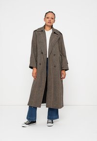 Long plaid coat in brown, black, and white hues, featuring a double-breasted front, wide lapels, and a relaxed fit. Worn over a white top and blue flared pants.