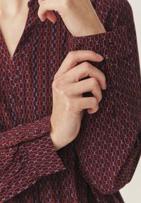 Burgundia pluus geomeetrilise kettmustriga punase ja sinise tooniga, nööbitava manseti ja kaeluses lipiku külge. Siledat kangast tekstuur.