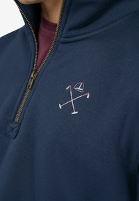 Suéter de marina con media cremallera, que presenta una textura suave. Logo bordado con palos de golf cruzados y una gorra en rosa claro y blanco.