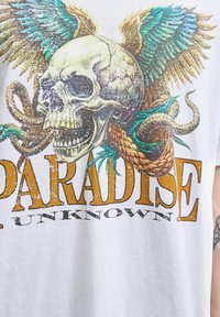Hvid grafisk t-shirt med et detaljeret kranie med vinger, slanger og teksten "PARADISE UNKNOWN" i orange, med teksturerede trykeffekter.