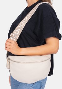 Kleine Umhängetasche aus Leder in hellem Beige mit einer geschwungenen Form. Sie verfügt über eine strukturierte Oberfläche, einen Reißverschluss und einen dekorativen, gemusterten Schulterriemen.