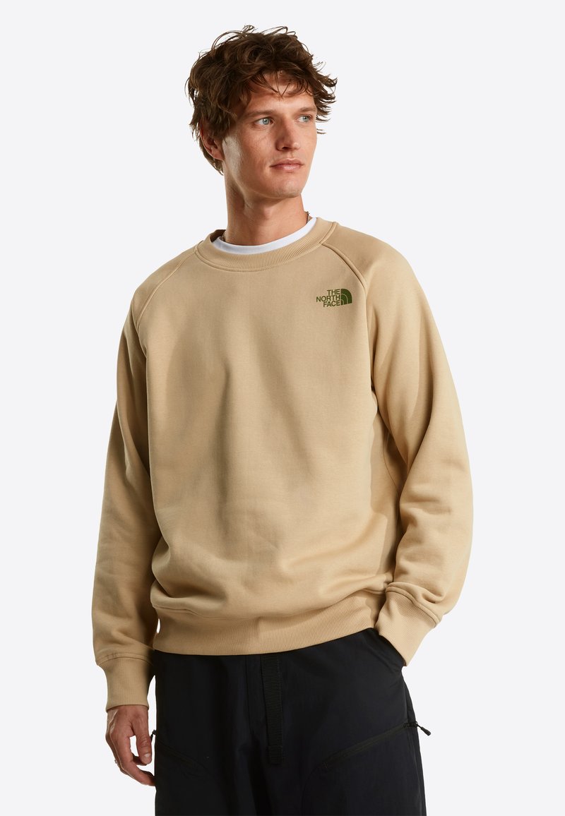 Young man wearing a beige The North Face sweatshirt and black pants, looking slightly to the right with a neutral expression.