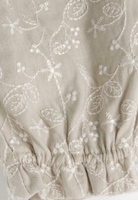 Tissu beige avec une broderie florale blanche complexe et des accents en relief. Détail de l'ourlet élastique froncé qui rehausse la texture et la forme.