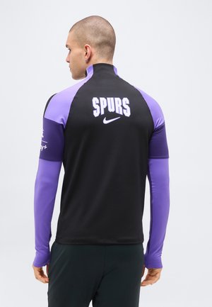 Mand med ryggen til iført sort og lilla Nike sportjakke med "SPURS" trykt på ryggen mod en ensfarvet baggrund.
