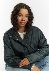 Giacca a vento verde scuro, con zip frontale, bottoni a pressione e vestibilità rilassata. Abbinata a una maglietta a righe, jeans in denim e capelli ricci.