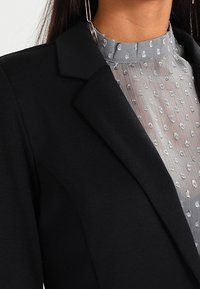 Svart kavaj med skarpa slag över en grå, genomskinlig blus med små vita prickar. Tygkvaliteten verkar slät med texturerade accenter.