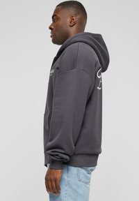 Charcoal grey hooded sweatshirt, cotton blend, featuring a front kangaroo pocket and ribbed cuffs, with logo print details on the sleeve.