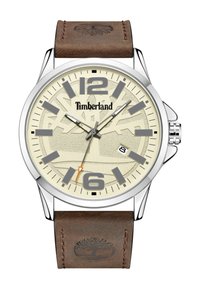 Reloj con correa de cuero marrón, caja redonda de plata, esfera color crema, marcadores de hora grises y manecilla de segundos naranja, presenta el logo de Timberland.