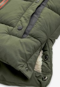 Chaqueta acolchada verde oliva con un suave forro blanco, que cuenta con botones y cremallera negros, junto con acentos de tela negra.