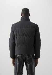 Sort polstret jakke med høj krave, der har et segmenteret quiltet design og elastiske manchetter. Materialet virker mat og struktureret.