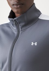 Pelēka ar rāvējslēdzēju aizdara jaka, kas izgatavota no gludas auduma, ar augstu apkakli un baltām plecu akcentiem, ar Under Armour baltu logotipu.
