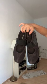 Črne tkaninske čevlje z elastičnim trakom čez nart, okroglo oblikovano konico in gladko teksturo, ki jih drži roka ob nevtralni ozadju.
