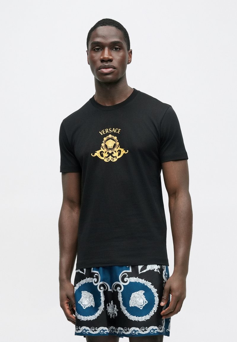 Sort bomuld T-shirt med guld "Versace"-logo og dekorativt design på brystet. Kortærmet og rund hals.