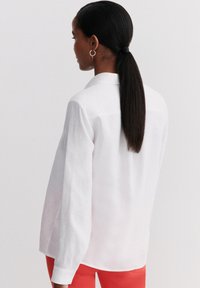 Camicia di lino bianca con maniche lunghe, colletto, vestibilità relaxed e texture liscia, indossata con pantaloni rossi brillanti.