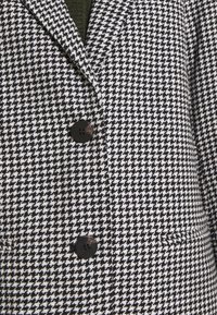 Blazer à motif pied-de-poule noir et blanc, avec un col cranté, deux boutons noirs et une coupe structurée avec poches.
