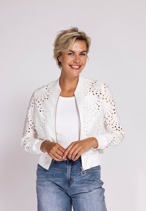 Lächelnde Frau mit kurzen blonden Haaren, die eine weiße Eyelet-Bomberjacke über einem weißen Top und blauen Jeans trägt, Hände vor dem Körper verschränkt.