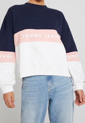Person trägt einen color-blocked Sweatshirt in Navy, Pink und Weiß mit dem Aufdruck „TOMMY JEANS“, kombiniert mit hellblauen Jeans aus Denim.