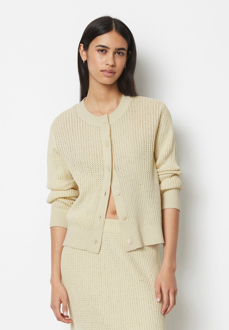 Knit cardigan in pale beige, featuring a textured pattern, round neckline, and button closure. Cuffs and hem are ribbed, providing a fitted look.