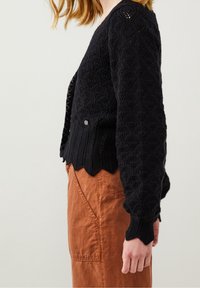 Czarny dziergany sweter z teksturowanym wzorem diamentowym, falistym dołem i srebrnym guzikiem po boku; noszony na rdzawych spodniach.