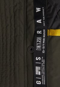 Olivengroen doorgestikt stof met gestikte lijnen, voorzien van een zwart etiket met witte tekst en een gele bandaccent. Inclusief een zwarte drukknoop.