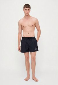 Young man standing barefoot wearing black EA7 branded swim shorts with elastic waistband and drawstring, hands relaxed by sides.