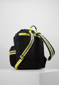Zaino in rete nera con spallacci e dettagli giallo brillante. Dotato di una maniglia superiore e cinturini laterali regolabili per una vestibilità sicura.