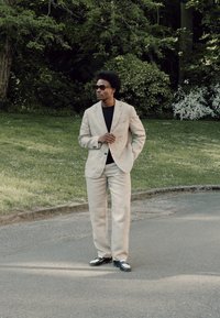Homme portant un costume beige, une chemise noire et des lunettes de soleil, debout sur un chemin pavé près de buissons verts et d’herbe dans un parc.