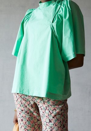 Vrouw draagt een losse lichtgroene blouse met pofmouwen en een broek met roze bloemenpatroon, ze houdt een kleine beige tas vast.