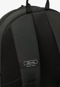 Sac à dos en tissu noir avec des bretelles rembourrées, une poignée supérieure et une étiquette visible avec "Superdry" et "OXFORD STREET" en blanc.
