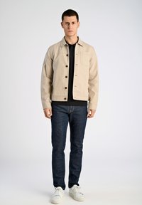 Giacca in cotone beige chiaro con bottonatura frontale, un taschino sul petto e bottoni marroni. Indossata sopra un maglione nero e jeans scuri.