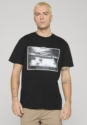 Młody mężczyzna z krótkimi blond włosami, ubrany w czarny t-shirt z białym napisem i wizerunkiem dymu, beżowe spodnie oraz srebrne kolczyki-koła.