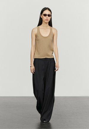 Vrouw met lang zwart haar die een donkere zonnebril draagt, een mouwloos beige topje, brede zwarte broek, lopend op een vlakke vloer tegen een lichte muur.