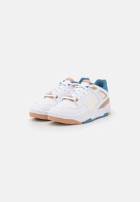 White sneakers with a smooth leather upper, light tan accents, blue tongue, and patterned rubber sole feature perforations for breathability.