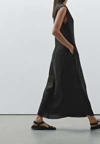 Musta, ilman hihoja oleva maksimekko, jossa on sivutaskut ja joka on valmistettu sileästä kankaasta. Yhdistettynä litteisiin mustiin sandaaleihin, joissa on naruistu.