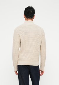 Beige strikkegenser med høy krage, med et teksturert mønster og ribbestrikkede mansjetter og hem. Modellen er vendt bort, noe som viser bakdetaljene.