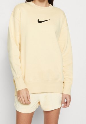 Žena v krémově zbarvené mikině Nike s černým logem, doplněná o odpovídající kraťasy, stojící před jednoduchým světlým pozadím.