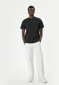 Sort t-shirt med korte ærmer parret med hvide cargo-bukser, der har sidelommer. Model iført hvide sneakers, stående foran en ensfarvet baggrund.