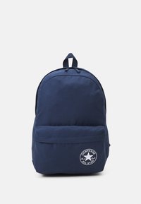 Mochila azul marino fabricada en tela, con la parte superior curva, un bolsillo frontal con cremallera y un logo blanco circular. Presenta dos correas para los hombros y un asa en la parte superior.