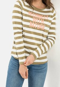 Personne portant un t-shirt à manches longues rayé olive et blanc avec le texte rose « RECONNECTER AVEC LA NATURE », associé à un jean bleu, les mains ajustant la manche.