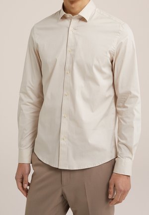 Mann trägt ein cremefarbenes langärmliges Hemd, das in hellbraune Hose gesteckt ist, mit entspannt an den Seiten liegenden Händen.