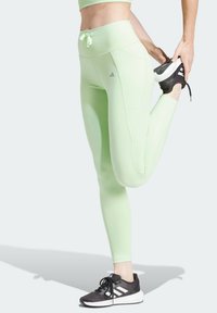 Persona che indossa leggings verde chiaro e sneaker neri Adidas si allunga i quadricipiti tirando un piede verso i glutei.