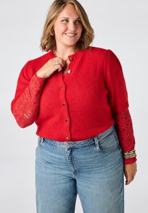 Lachende vrouw met schouderlang blond haar draagt een rood vestje met knopen, lange kanten mouwen, blauwe spijkerbroek en gouden armbanden om haar pols.