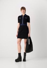 Robe noire à manches courtes, col en denim et poches, détail de fermeture éclair argentée, associée à des bottes compensées noires et un sac plissé.