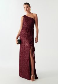Jednoduché jednoramenné vínově červené šaty pokryté flitry s vysokým rozparkem a volánkovým detailem v pase. Modelka drží černou kabelku a má na sobě metalické sandály.