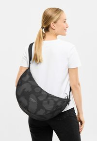 Čierna vzorovaná crossbody taška s polmesiacovým tvarom, vyrobená z textílie, s hladkou textúrou a nastaviteľným popruhom. Viditeľné logo.