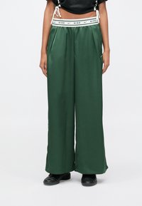 Κάτω μέρος σώματος ενός ατόμου που φοράει σκούρο πράσινο φαρδύ παντελόνι Nike με λευκή μέση και μαύρα αθλητικά παπούτσια σε λευκό φόντο.