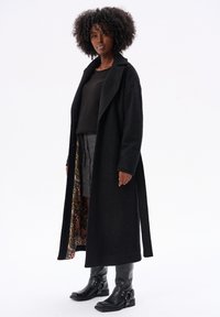 Czarny długi płaszcz o teksturowanym wykończeniu, z podszewką w kwiatowy wzór, zestawiony z ciemnym swetrem i spódnicą w kratę do kolan.